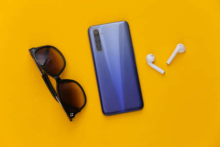 Modern accessories. Modern smartphone with wireless earphones, sunglasses on yellow background. top...