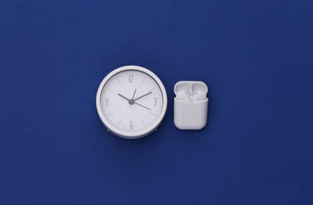 White clock and wireless earphones in the charging case on classic blue background. top viewの写真素材