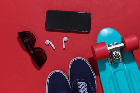 Cruiser board, smartphone and wireless headphones, sunglasses and sneakers on bright red background....
