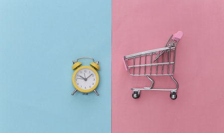 Mini supermarket trolley and retro alarm clock on pink blue pastel background. shopping time. top...
