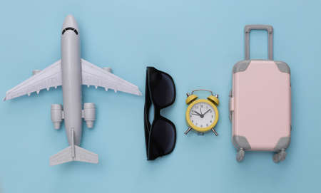 Air plane, mini travel luggage, sunglasses and alarm clock on blue background. Time to travel. top...