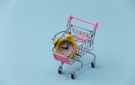 Yellow retro alarm clock in supermarket trolley on blue background.