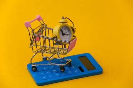 Yellow retro alarm clock in supermarket trolley and calculator on yellow background. Savings, shopping costsの写真素材
