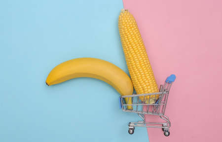 Mini Supermarket trolley with swing of corn and banana on pink blue pastel background. Minimalist food flat lay. top viewの写真素材