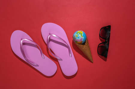 Flip flops, ice cream waffle cone with globe, sunglasses on red background. Summer flat lay. top viewの写真素材
