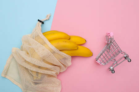 Eco cotton bag with ripe bananas and mini shopping trolley on pink blue pastel background. top view. flat layの写真素材
