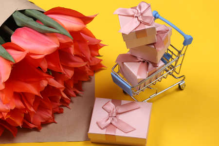 Bouquet of tulips and shopping trolley with gift boxes on yellow background. holiday shopping. Mother's Day or March 8th, birthday.の写真素材