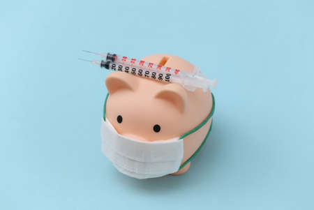 Piggy bank in medical mask with syringes on blue background. vaccination. economic disease. financial crisis. Covid-19 pandemicの写真素材