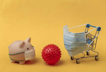 Mini supermarket trolley and piggy bank with medical mask, virus strain model on yellow background. Covid-19 pandemicの写真素材