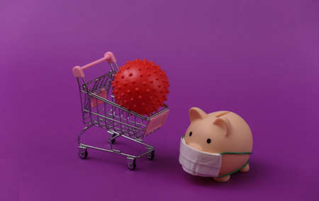 Piggy bank in a medical mask with a shopping trolley, virus strain model on purple background. Shopping during covid-19, economic crisis, economyの写真素材