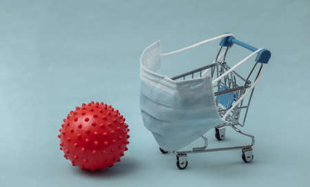 Mini supermarket trolley with medical mask, virus strain model on blue background. Shopping during...