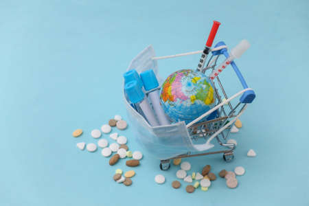 Shopping trolley in medical mask with test tubes, syringes, pills and a globe on a blue background. Global healthcareの写真素材