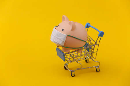 Piggy bank in a medical mask with a shopping trolley on yellow background. Shopping during covid-19, economic crisis, economyの写真素材