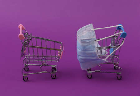 Mini supermarket trolleys with medical mask on purple background. Shopping during the Covid-19...