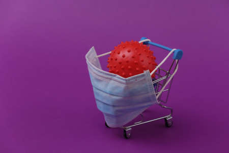 Mini supermarket trolleys with medical mask, virus strain model on purple background. Shopping during the Covid-19 pandemicの写真素材