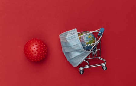Mini supermarket trolley with medical mask, virus strain model, globe on red background. Shopping during the Covid-19 pandemicの写真素材