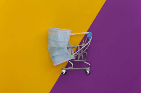 Mini supermarket trolley with medical mask on purple yellow background. top view. flat layの写真素材