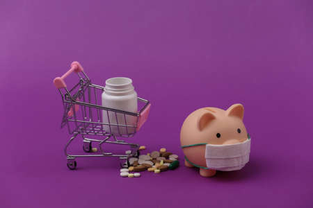 Piggy bank in a medical mask and shopping trolley with pills bottle on purple background.の写真素材