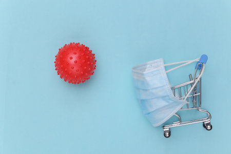 Mini supermarket trolley with medical mask, virus strain model on blue background. Shopping during the pandemic. top viewの写真素材