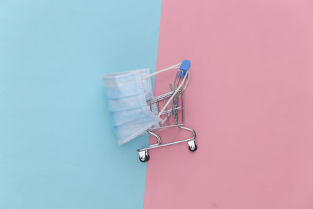 Mini supermarket trolley with medical mask on pink blue pastel background. top viewの写真素材