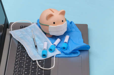 Piggy bank in medical mask, test tubes, gloves with syringe on laptop keyboard.の写真素材
