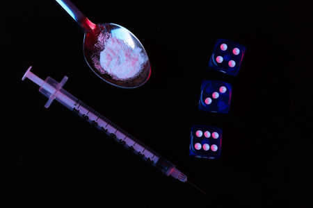 Drug and gambling addiction. Spoon with a drug powder, syringe, dice on a black background with red-blue neon lightの写真素材