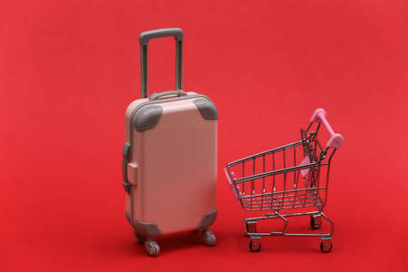 Travel and shopping concept. Plastic travel suitcase and supermarket trolley on red background.