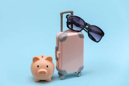 Travel or trip concept. Plastic travel suitcase with sunglasses, piggy bank on blue background. Minimal styleの写真素材