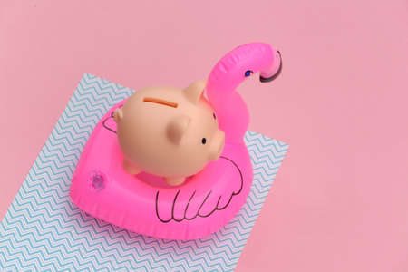 Inflatable pink flamingo with piggy bank on pink blue pastel background. Summer vacation concept....
