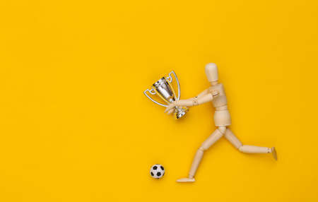Wooden puppet runs with soccer ball and championship cup on yellow backgroundの写真素材
