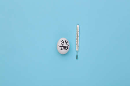 The egg face in a medical mask and thermometer on blue background. Covid-19 pandemic. top view