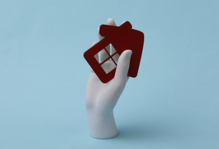 White mannequin hand holds a house figurine on blue background. Buying propertyの写真素材