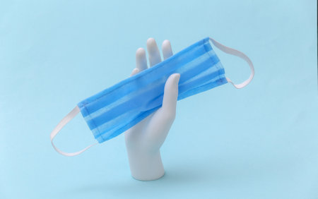 A white mannequin hand holds a medical mask on a blue background. medicine minimalism concept