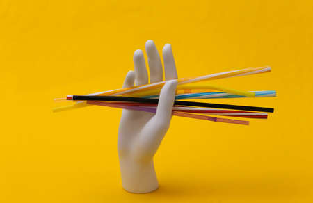 White mannequin hand holding many plastic cocktail tubes on yellow background
