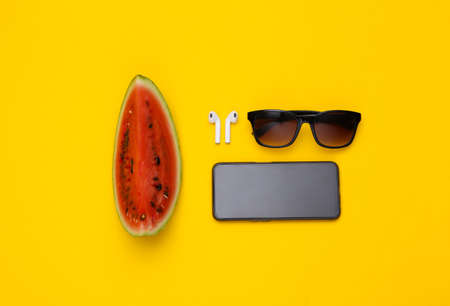 Slice of ripe watermelon, wireless headphones, sunglasses and smartpone on yellow background. summer fun. top view. flat layの写真素材