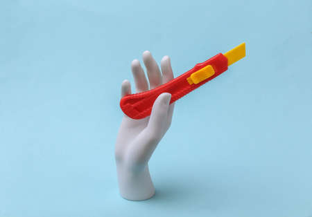 White mannequin hand holds toy paper knife on blue background. Renovation concept, industry....