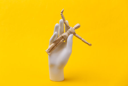 White mannequin hand holds wooden puppet on yellow backgroundの写真素材