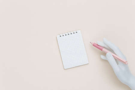 mannequin hand holds pen and notepad on beige background. top view