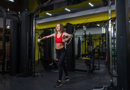 Smilling Fit woman flexing muscles on gym machine. Girl perform exercise with exercise-machine Cable Crossover in modern gym. fitness and bodybuildingの写真素材