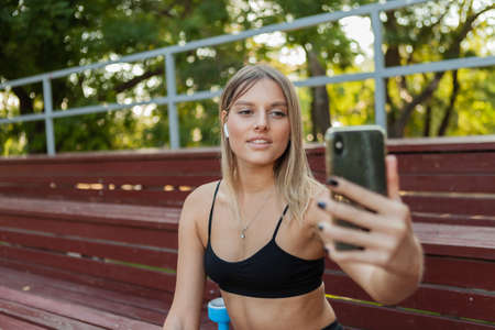Young attractive woman in sportswear resting while sitting on wooden stands after morning outdoor workout and doing selfie on smartphoneの写真素材