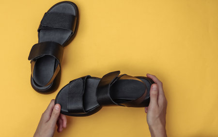 Female hands hold leather black fashionable sandals on yellow background. top viewの写真素材