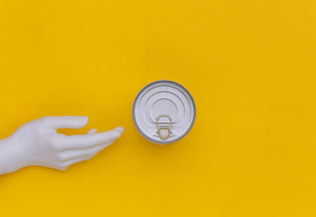 Metal can of canned food and mannequin hand on yellow background. minimalism. top view