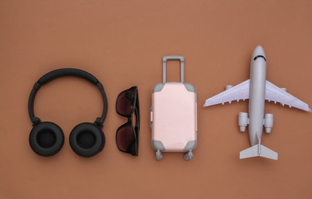 Mini travel luggage with stereo headphones, sunglasses and air plane on brown background. travel...