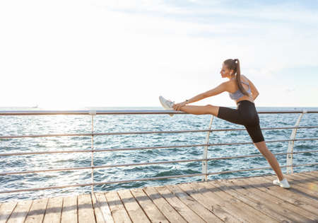 Slim fit woman with perfect body is practicing leg stretching before training on the beach at...