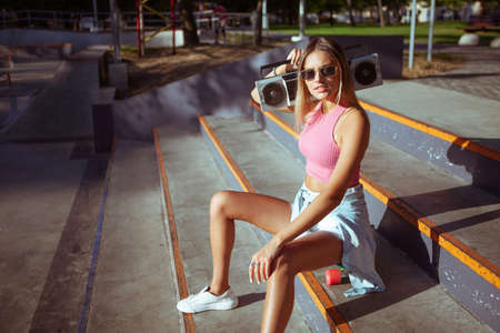 Funny stylish young woman skater with cruiser board and retro tape recorder sits on stairs in skateparkの写真素材