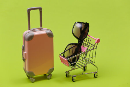 Travel and shopping concept. Mini plastic travel suitcase and supermarket trolley with sunglasses on green background.の写真素材