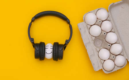 Chicken egg happy face with headphones and eggs tray on a yellow background. top view. flat layの写真素材