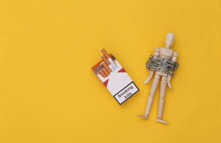 smoking addiction. Wooden puppet wrapped in steel chain and pack of cigarettes on yellow background