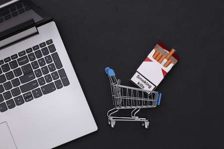 Laptop and shopping trolley with pack of cigarettes on black background. top view