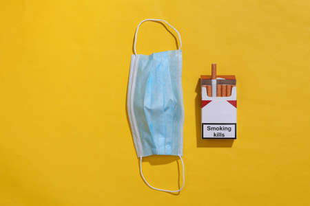 Pack of cigarettes and medical face mask on yellow background. top view